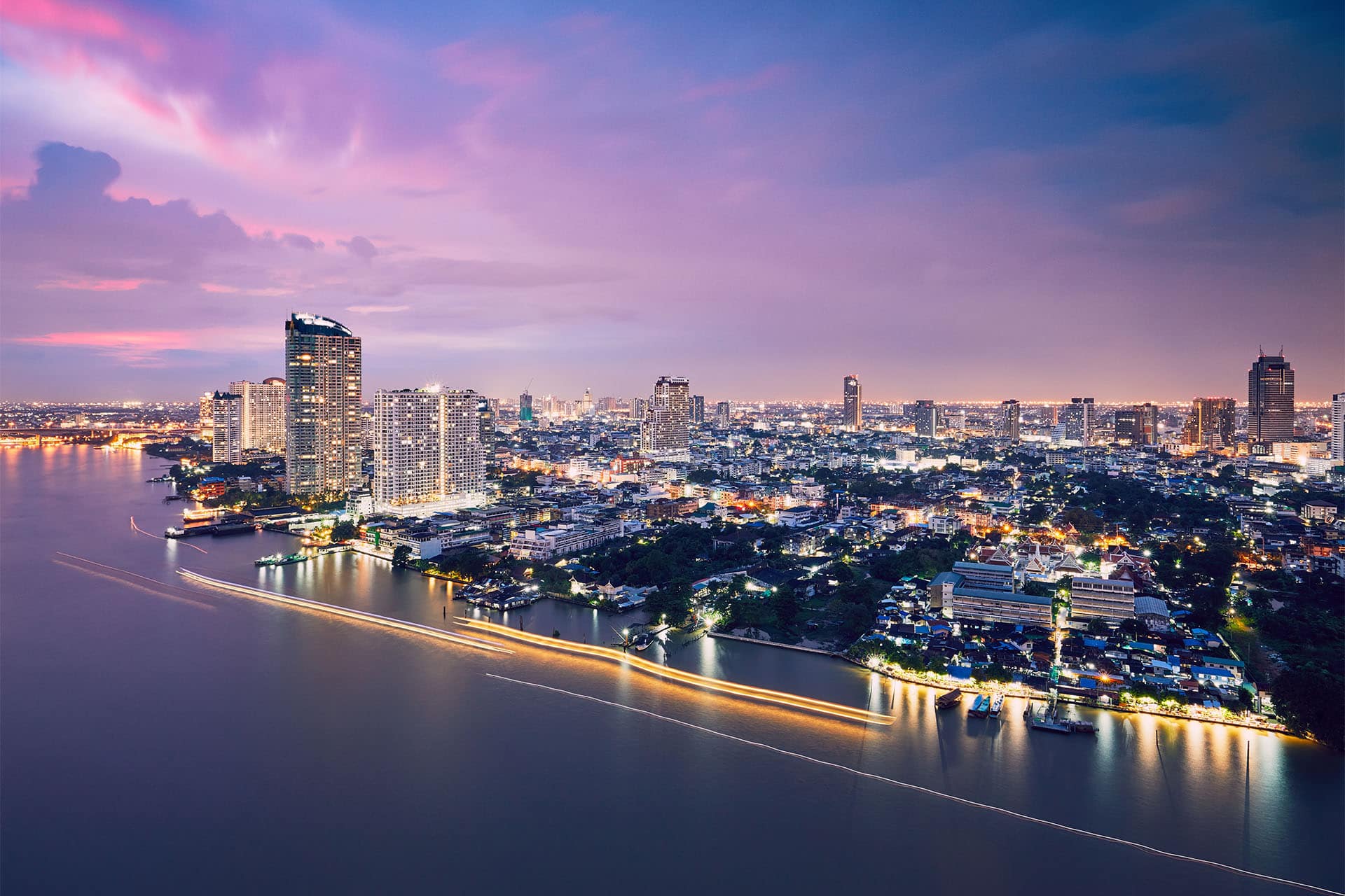 Luxury riverside condominiums and high-rise buildings in Bangkok overlooking the Chao Phraya River with city lights glowing at dusk.