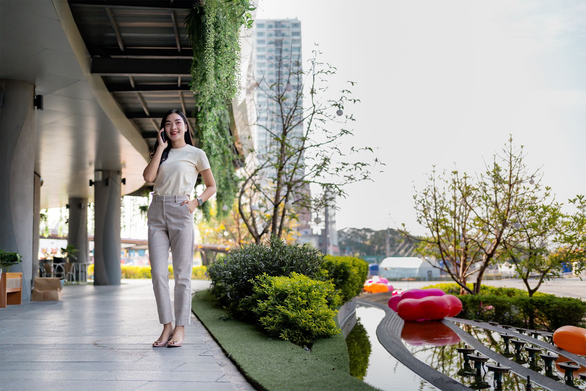 Young woman standing and talking on the phone in a modern Bangkok residential complex with landscaped greenery and outdoor seating.